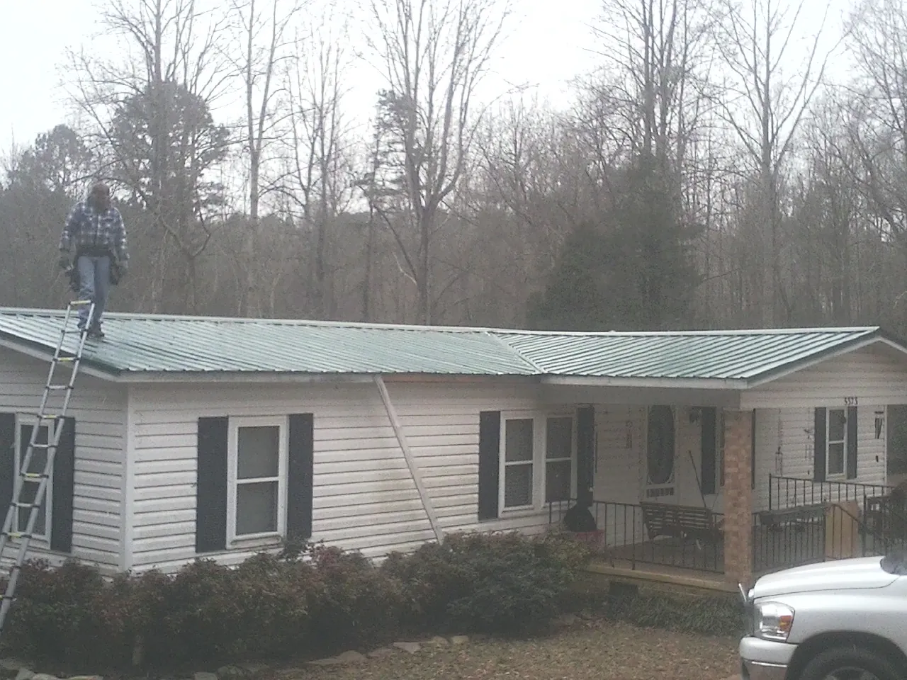 Worker on a ladder beside a new green metal roof on a ranch home in Prairie Grove
