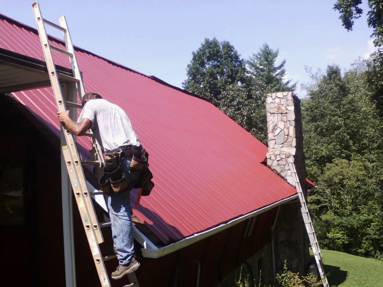 Roofer on a ladder beside a red metal roof and stone chimney — Soffit & Fascia Repair by Roofing Brothers in Prairie Grove